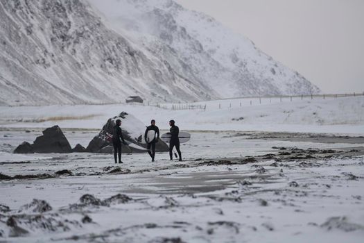 Authentic local Arctic surfers going by beach after surfing in Norwegian sea at snow capped mountain background in winter. Unstad, Norwegian village on Lofoten islands. Norwegian sea coastline. Winter water activities extreme sport