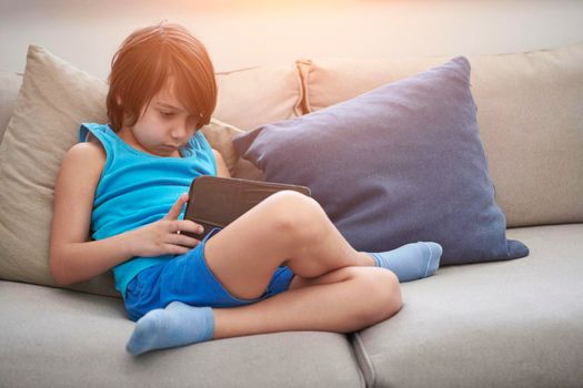 Little Arabian boy sitting on sofa and playing game on digital tablet. Portrait of a young arab child at home watching cartoon on the laptop. Modern kid and online education elearning technology.