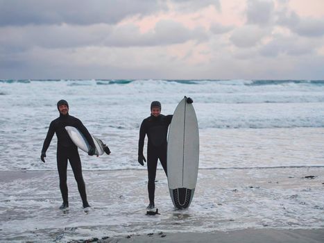 Authentic local Arctic surfers going by snowy beach after surfing in Northern sea. Norwegian sea coastline. Winter water activities extreme sport