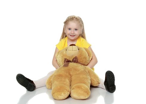 A lovely little round-faced blonde girl, with very long beautiful hair, in short skirts and yellow jerseys.She hugs a big teddy bear.Isolated on white background.