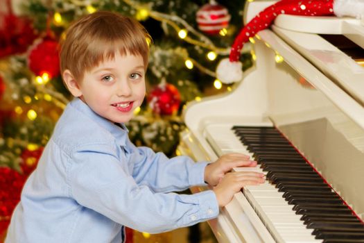 A little boy is sitting at the piano near the Christmas tree. Family holidays concept.