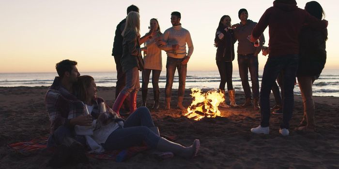 Happy Carefree Young Friends Having Fun And Drinking Beer By Bonefire On The Beach As The Sun Begins To Set