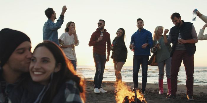 Happy Carefree Young Friends Having Fun And Drinking Beer By Bonefire On The Beach As The Sun Begins To Set