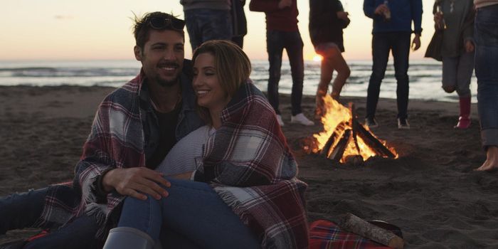 Happy Carefree Young Friends Having Fun And Drinking Beer By Bonefire On The Beach As The Sun Begins To Set