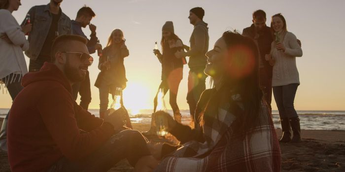 Happy Carefree Young Friends Having Fun And Drinking Beer By Bonefire On The Beach As The Sun Begins To Set