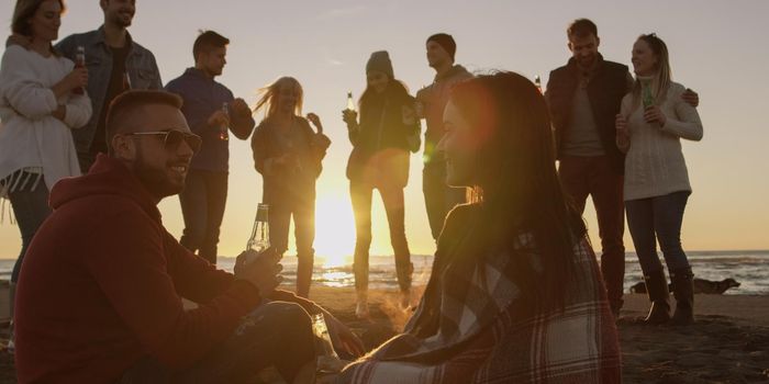 Happy Carefree Young Friends Having Fun And Drinking Beer By Bonefire On The Beach As The Sun Begins To Set