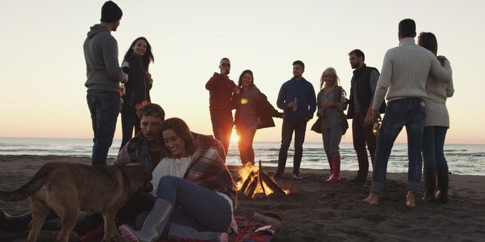 Happy Carefree Young Friends Having Fun And Drinking Beer By Bonefire On The Beach As The Sun Begins To Set