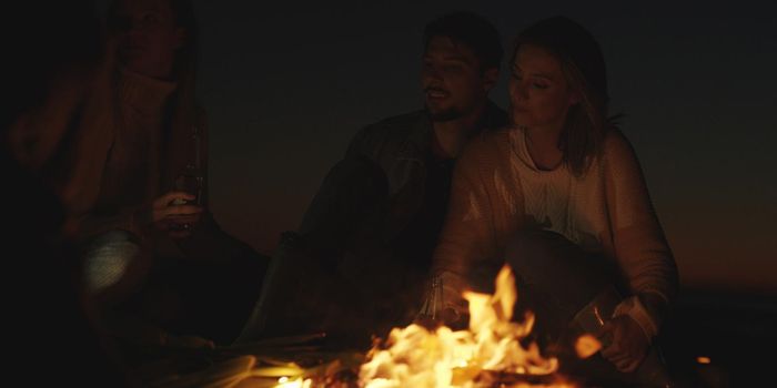 Young Couple Sitting with friends Around Campfire on The Beach At Night drinking beer