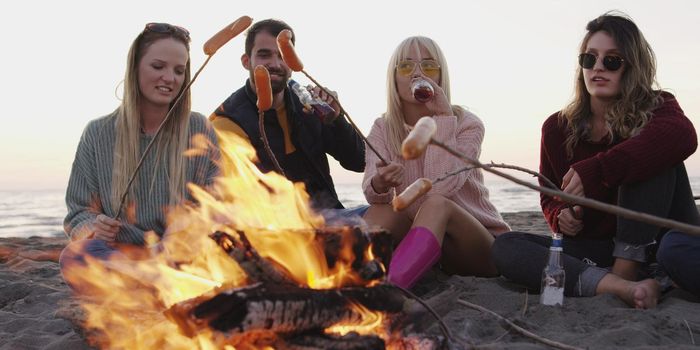 Group of young friends sitting by the fire late at night, grilling sausages and drinking beer, talking and having fun