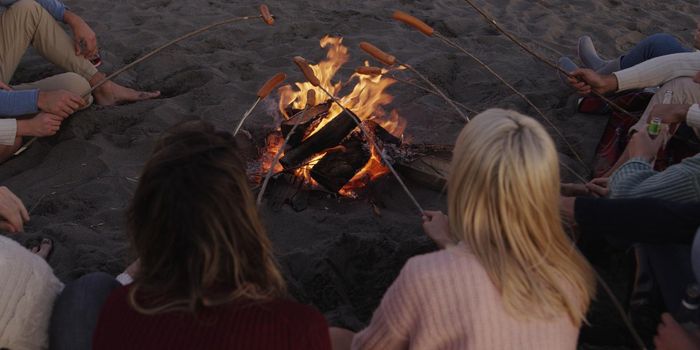 Group of young friends sitting by the fire late at night, grilling sausages and drinking beer, talking and having fun