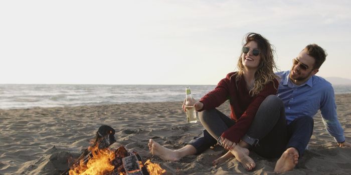 Young Couple Relaxing By The Fire, Drinking A Beer Or A Drink From The Bottle.