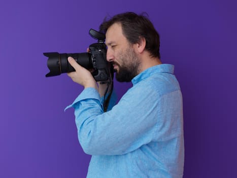 Portrait of  male Photographer  with beard in blue shirt holding digital dslr  camera while standing against purple  background