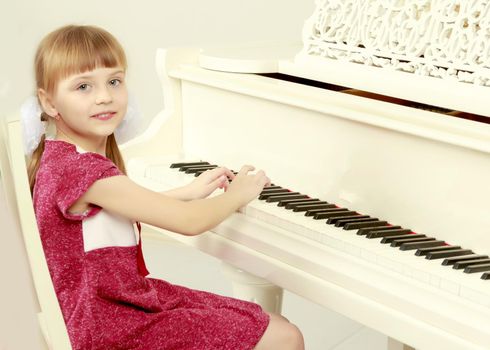 Beautiful little blonde girl with short bangs and pigtails on her head in a good mood.She sits opposite the white piano and tries to play it.