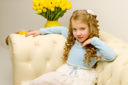 Beautiful little girl with long silky hair, sits on the couch with a bouquet of yellow tulips. The concept of happy people, style and fashion.