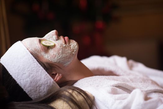 Beautiful young woman is getting facial clay mask at spa, lying with cucumbers on eyes