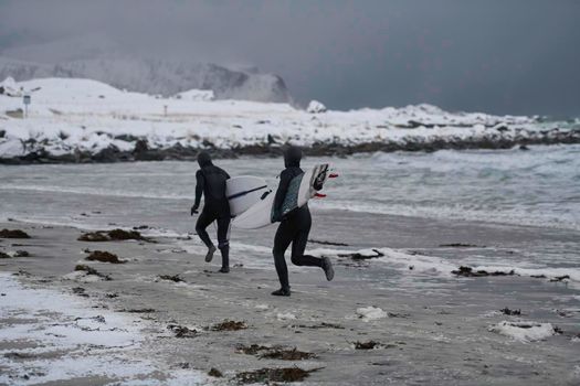 Authentic local Arctic surfers running on snowy  beach after surfing in Norwegian sea. Snow capped mountain background in winter. Norwegian Lofoten islands. Norwegian sea coastline. Winter water activities extreme sport