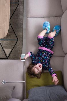 top view of cute little girl using a smartphone and smiling while lying on the sofa at home