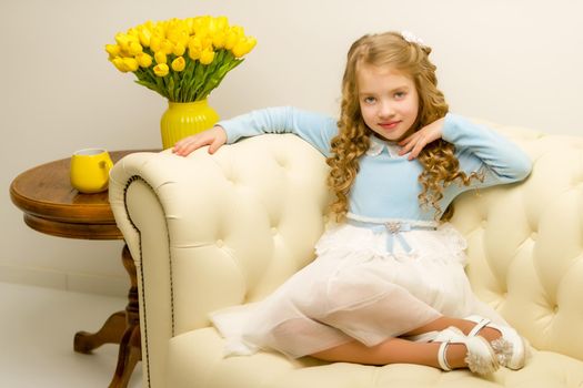 Beautiful little girl with long silky hair, sits on the couch with a bouquet of yellow tulips. The concept of happy people, style and fashion.