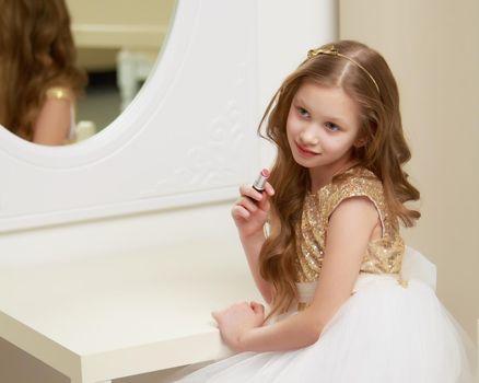 A nice little girl near the big mirror paints her lips with lipstick. The concept of childhood, style and fashion.