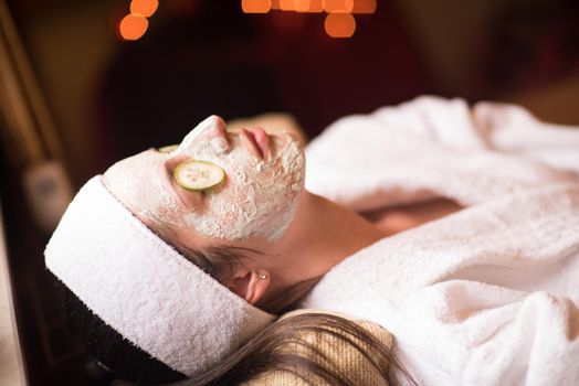 Beautiful young woman is getting facial clay mask at spa, lying with cucumbers on eyes