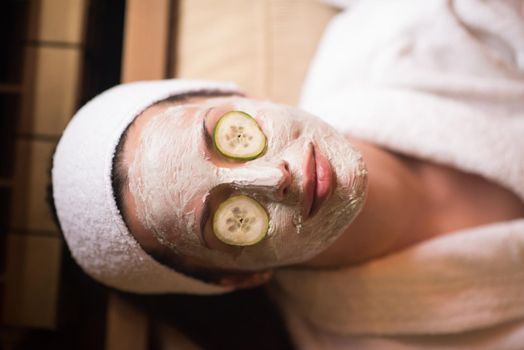 Beautiful young woman is getting facial clay mask at spa, lying with cucumbers on eyes