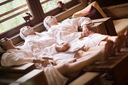 beautiful young women  getting facial clay mask at spa, lying with cucumbers on eyes