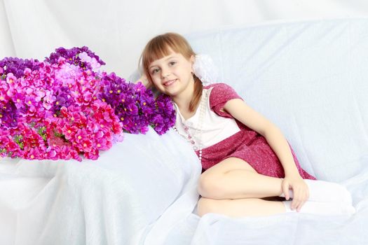 Beautiful little blonde girl with short bangs and pigtails on her head in a good mood.She sits next to a bouquet of flowers.