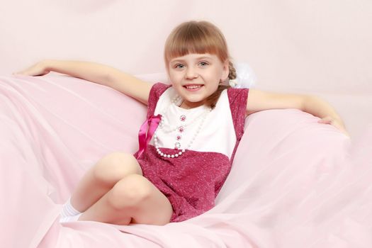 Beautiful little blonde girl with short bangs and pigtails on her head in a good mood.She sits on a pink sofa.