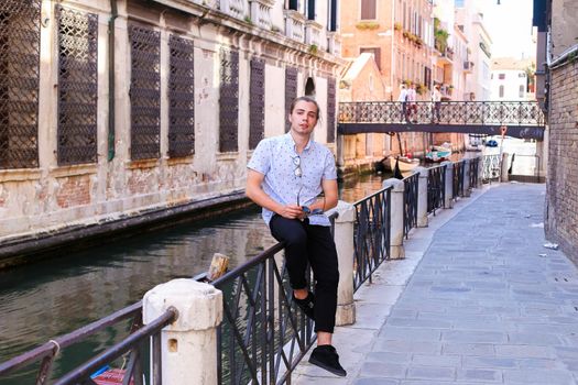 Young male tourist sitting on banister in Venice, Italy. Concept of last minute tours to Europe and romantic venetsian street.