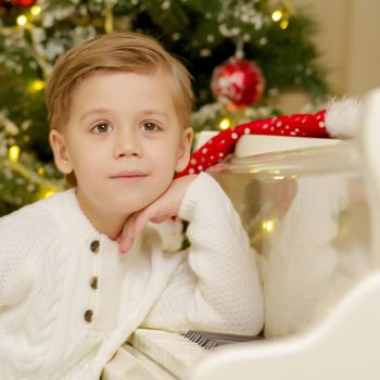 A little boy is sitting at the piano near the Christmas tree. Family holidays concept.