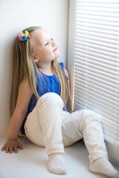 Cute little girl with long blond hair below the shoulders. In the blue shirt and white jeans sitting on the windowsill. Girl looking out the window
