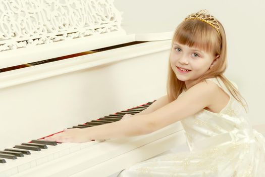 A lovely little girl with long, luxurious, blond hair in a white ball gown.A girl is posing near a white grand piano.