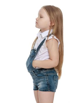 Offended little chubby girl with long blonde hair below the waist, short denim overalls. The girl turned away from the camera and turned sideways. Close-up