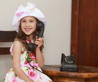 Cheerful little girl in a white dress with roses and white hat, calling on the old phone. Girl sitting on an old Viennese chair at the old round table . Retro style . close-up