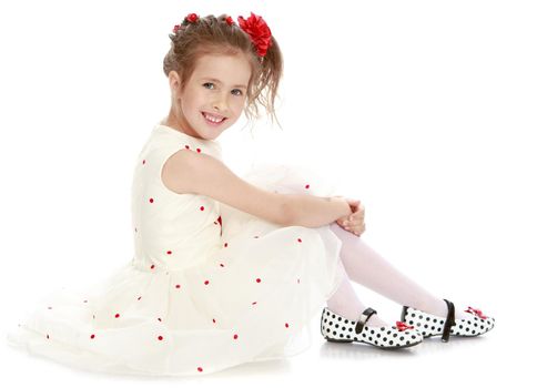 Nice little Princess dressed in a white dress with a red flower on her head . Girl sitting on the floor turned sideways to the camera - Isolated on white background