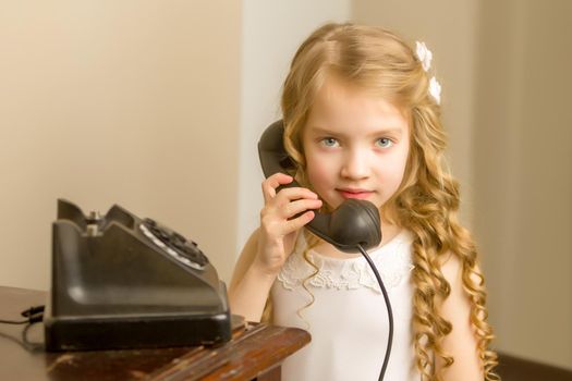 A beautiful little girl in a room in the fifties of the last century, talking on an old phone with a big black tube. Retro style, black and white photo in the studio. Concept of nostalgia, vintage.