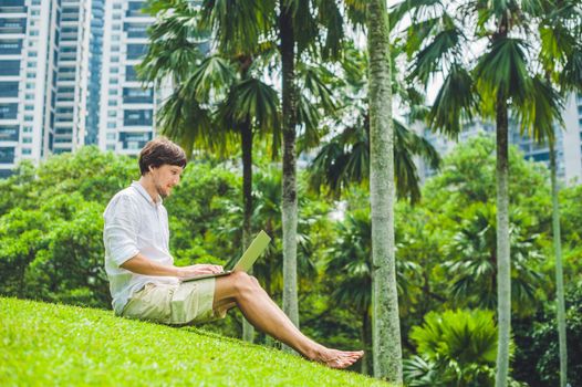 Man businessman or student in casual dress using laptop in a tropical park on the background of skyscrapers. Dressing in a white shirt, beige shorts. Mobile Office concept.