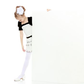 Beautiful little blonde girl dressed in a white short dress with black sleeves and a black belt.The girl peeks out from behind white banner.Isolated on white background.