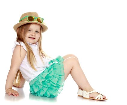 Caucasian little girl with long blond hair below the shoulders , a hat and sunglasses and a short green skirt . Girl sitting on the floor turned sideways to the camera - Isolated on white background
