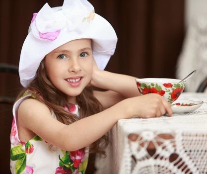 Gentle little girl in a white dress with roses and white hat drinking tea. Girl sitting on an old Viennese chair behind the old oak table , covered with a lace tablecloth. Retro style . close-up