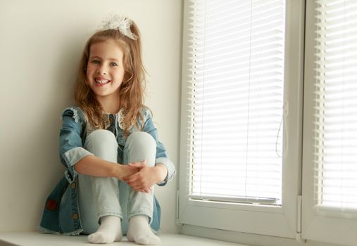 Cute little girl sitting on the windowsill near the window , around the closed blinds. The girl hugged her knees