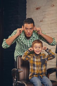 stylish little kid sitting on chair at barbershop with his young father on background, they are fooling around and having fun