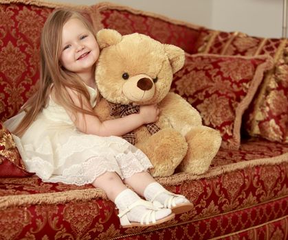 Adorable little round-faced girl with long, blonde hair below the shoulders, little Princess in a white dress. Girl hugging Teddy bear