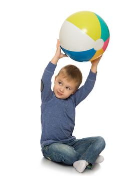 Beautiful little boy in jeans and a blue shirt. The boy sits on the floor with the ball . He raised the ball above his head - Isolated on white background