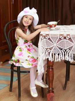 Gentle little girl in a white dress with roses and white hat drinking tea. Girl sitting on an old Viennese chair behind the old oak table , covered with a lace tablecloth. Retro style