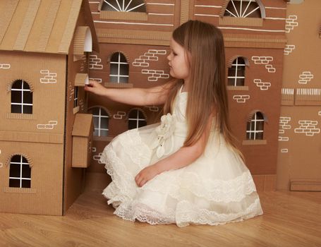 Beautiful little girl with long blond hair below the shoulders , in white dress plays with a doll's house