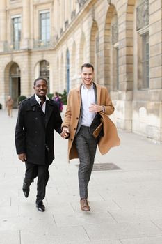 Caucasian european man running with afroamerican male person and holding hands in city. Concept of happy gays and strolling.