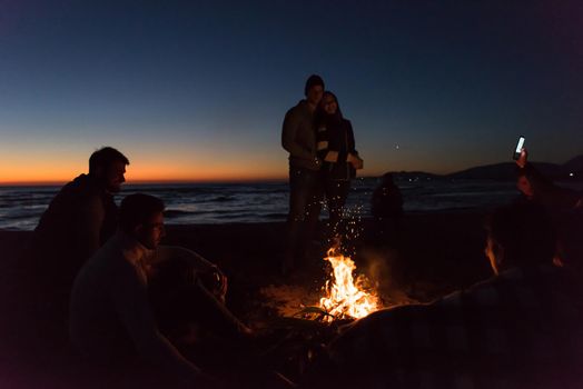 Happy Carefree Young Friends Having Fun And Drinking Beer By Bonefire On The Beach As The Sun Begins To Set