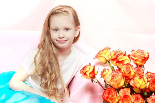 A beautiful little girl with long, light, curly hair, in a blue long skirt. She sits on a pink couch beside a large bouquet of tea roses.