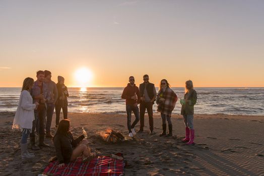 Happy Carefree Young Friends Having Fun And Drinking Beer By Bonefire On The Beach As The Sun Begins To Set